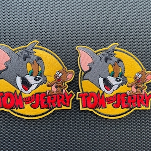 May include: Two embroidered iron-on patches featuring Tom and Jerry. The patches are yellow with red and black lettering that says "Tom and Jerry". Tom is a gray cat with a mischievous grin and Jerry is a brown mouse.