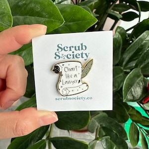 May include: A gold-colored enamel pin featuring a scroll with the text "Chart like a Lawyer" and a feather quill. The pin is on a white card with the "Scrub Society" logo and website address. The background is a lush green plant.