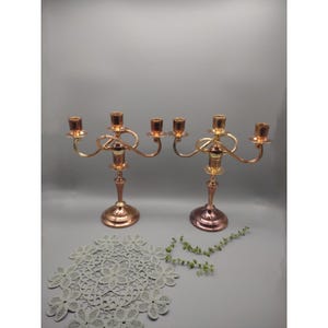 May include: A pair of gold and copper colored metal candle holders with three arms each. The candle holders are on a white lace doily.
