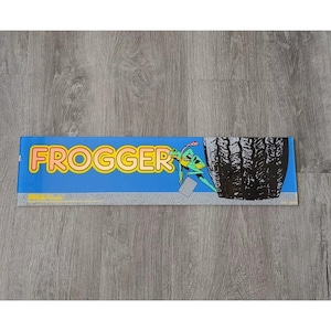 May include: A blue and yellow arcade game marquee with the word "FROGGER" in large, yellow letters. A green frog is climbing a tire.