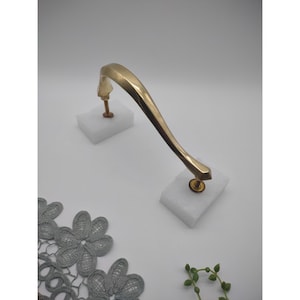 May include: A gold-coloured door handle with a curved design. The handle is mounted on two white foam blocks. The handle has a decorative, geometric end. A grey lace doily and a sprig of greenery are in the foreground.