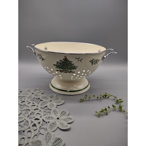 May include: A white enamel colander with a green and red Christmas tree design. The colander has a silver rim and handles.