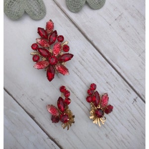 May include: A set of three red rhinestone jewelry pieces. A large brooch with a floral design and two smaller earrings with similar designs.