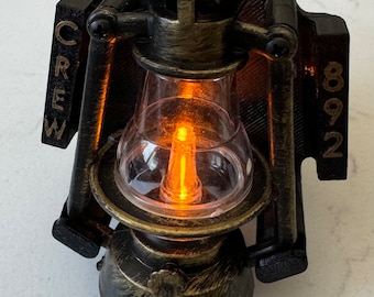 Puede incluir: Una linterna de metal negro y dorado con un globo de vidrio transparente y una bombilla encendida en su interior. La linterna tiene las palabras "CREW" y "892" en los lados.