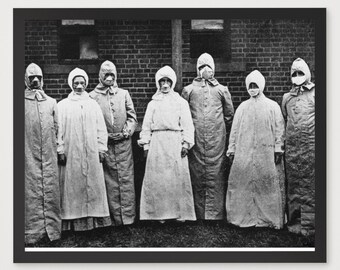 May include: Black and white framed print featuring a historical photograph of a group of people wearing protective suits and hoods. The subjects stand in front of a brick building. The image is mounted on a white mat.