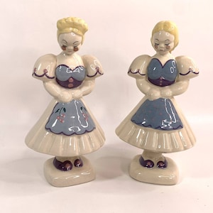 May include: Two ceramic figurines of women in blue and white dresses with yellow hair. The women are standing on white bases.
