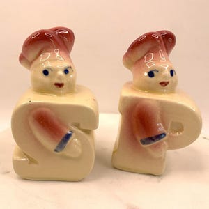 Op de afbeelding: Een paar vintage keramische zout- en peperstrooiers. Elke strooier is gevormd als een kok met een rode hoed en een crèmekleurig lichaam. De armen van de kok zijn beschilderd met rode en blauwe accenten.