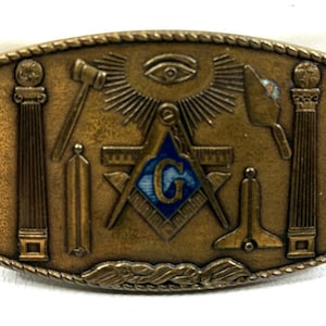 May include: A rectangular, antique-style belt buckle with Masonic symbols. The bronze-colored buckle features a central square and compass design with a blue 'G', surrounded by other emblems like a hammer, pillars, and an all-seeing eye.