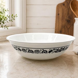 Może przedstawiać: Biała ceramiczna miska z czarnym kwiatowym wzorem wokół zewnętrznej krawędzi. Miska ma szeroki, okrągły otwór i gładkie, błyszczące wykończenie, prezentujące szczegółowy botaniczny wzór z kwiatami i liśćmi wykonany czarnym tuszem.