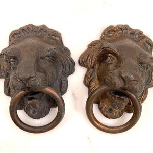 May include: A pair of antique bronze lion head door knockers. Each knocker depicts a detailed lion's face with a ring in its mouth. The bronze has a weathered look, with colour and texture variations, enhancing their vintage appeal.
