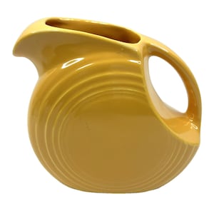 Puede incluir: Una jarra de cerámica de color amarillo brillante que presenta un cuerpo redondeado con detalles de anillos concéntricos en el lateral. El recipiente incluye un asa curva y un pico vertedor prominente en la parte superior, con acabado brillante.