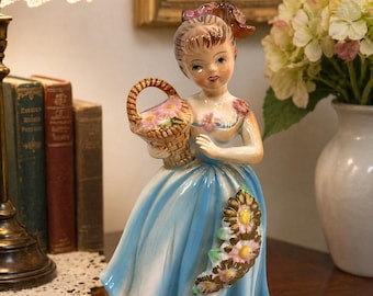 May include: A vintage porcelain figurine of a woman in a blue dress holding a basket of flowers. The figurine has a detailed face and is decorated with floral accents. The dress has a flowing design, and the figurine is displayed on a wooden surface.