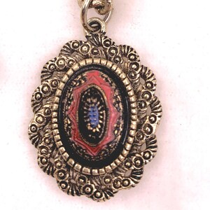 Könnte beinhalten: Vintage ovaler Anhänger mit goldfarbenem Filigranrahmen. Der Anhänger hat eine schwarze Mitte mit roten und blauen Akzenten. Die Kette ist ebenfalls goldfarben. Ein klassisches Schmuckstück.