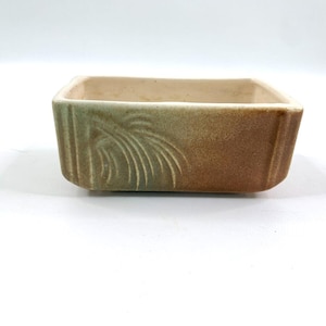 Puede incluir: Una jardinera rectangular de cerámica con una combinación de colores verde y marrón descoloridos. La jardinera presenta un diseño texturizado en un lado, con un patrón curvo en forma de hoja. El interior es de color beige claro.