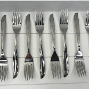 Puede incluir: Un juego de nueve tenedores de acero inoxidable dispuestos en una bandeja blanca. Los tenedores tienen un diseño elegante con un mango curvo y un pequeño detalle de estrella cerca de las púas. Ideales para la mesa diaria.