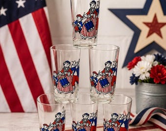 May include: A set of six clear glass tumblers stacked in a pyramid formation. Each glass features a colorful graphic print of a marching band with figures in red, white, and blue attire, including a drummer and a flute player in a patriotic style.