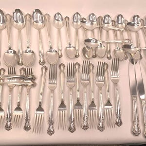 May include: A set of silver cutlery, including spoons, forks, and knives, arranged in a row on a white surface.