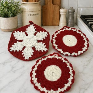 Op de afbeelding: Drie handgemaakte gehaakte pannenlappen. Eén is een bordeauxrode zeshoek met een wit sneeuwvlokontwerp. De andere twee zijn rond, bordeauxrood met witte geschulpte randen en centra. Keukendecoratie.