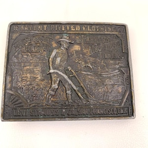 May include: A vintage brass belt buckle with a raised image of a farmer plowing a field. The buckle is embossed with the text "Patent riveted clothing, The Best in Use For Farmers, Mechanics & Miners, Levi Strauss & Co. San Francisco Cal."