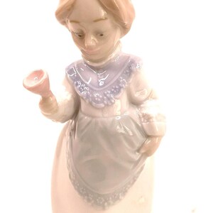May include: A porcelain figurine of a woman in a long dress, holding a pink object. The figurine has a light skin tone, with a light brown hairstyle and a light blue collar. The dress is white and light blue.