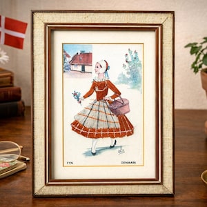 Könnte beinhalten: Gerahmter Aquarelldruck einer Frau in traditioneller dänischer Tracht, die einen Korb und Blumen trägt. Der Druck trägt die Aufschrift "FYn Denmark" und befindet sich in einem goldfarbenen Rahmen. Im Hintergrund ist eine dänische Flagge zu sehen.