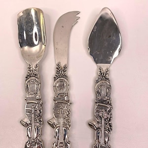 Può includere: Set di tre utensili da portata argentati. Il set include un cucchiaio, un coltello da formaggio e una paletta per torte. Ogni manico è finemente decorato con un motivo a violino, aggiungendo un tocco di eleganza. Gli utensili sono probabilmente in acciaio inossidabile.