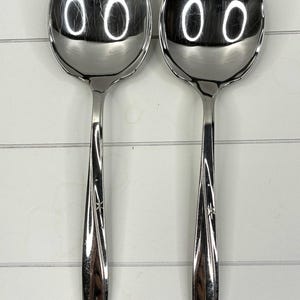 Puede incluir: Dos cucharas soperas de acero inoxidable con cuencos ovalados y mangos largos y decorativos. Los mangos tienen un diseño de estrella cerca de la base. Las cucharas son reflectantes.