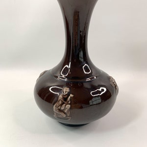 May include: A dark brown ceramic vase with a wide mouth and a narrow neck. The vase has a glossy finish and is decorated with raised figures of monkeys.
