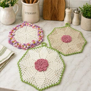 May include: Three handmade crochet pot holders in a kitchen setting. One is white with a pink and purple floral border, another is beige with a pink flower center, and the third is white with a green border and pink flower center.