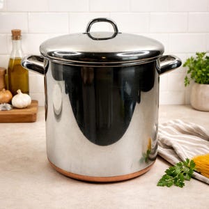May include: A large, stainless steel stockpot with a matching lid. The pot has two side handles and a copper-colored base. The lid features a single, arched handle. The pot is sitting on a light-colored countertop.