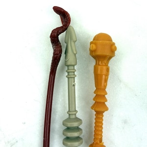 Puede incluir: Tres armas de juguete: un bastón en forma de serpiente rojo oscuro, una lanza gris claro y una maza naranja. El bastón de serpiente es curvo con una superficie texturizada. La lanza tiene una punta y una base decorativa. La maza tiene una parte superior redondeada y un mango acanalado.