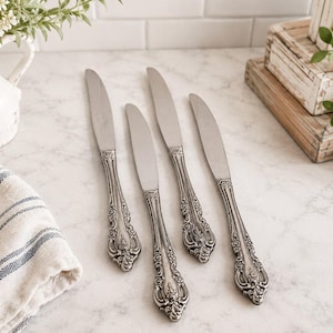 May include: A set of four silver-toned metal knives featuring ornate, embossed floral and scrollwork detailing on the handles. The blades are smooth and polished, while the handles exhibit a darkened, textured finish with intricate relief patterns.