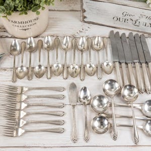 May include: A large set of silver-toned flatware including multiple forks, knives, and spoons of varying sizes. The collection features a consistent decorative pattern on the handles and includes several round-bowled soup spoons and a butter knife.