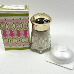 Pode incluir: Um decantador de pó de vidro com tampa de metal dourado com orifícios perfurados, acompanhado pela sua caixa retangular original rotulada como Charisma e Net Wt 1.25 oz, ao lado de uma pequena esponja aplicadora branca e redonda.