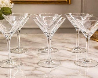 May include: Set of six clear glass martini glasses. Each glass has a conical bowl and a long stem. The glasses are arranged on a marble surface, with a vase of white flowers and a gold-colored decorative object in the background.