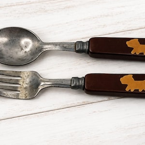 May include: Vintage child's spoon and fork set. The spoon and fork are made of metal with dark brown handles. Each handle has a light brown dog silhouette. The set is displayed on a white wooden surface.