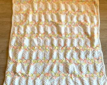May include: A handmade, white crochet blanket with a scalloped edge. The blanket features a chevron pattern with rows of yellow and peach accents. The blanket is rectangular and appears to be made of soft yarn.
