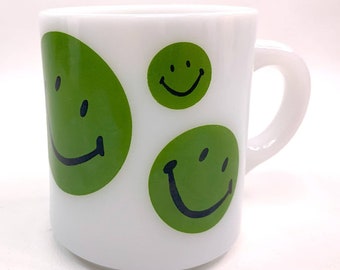May include: White ceramic mug with three green smiley faces. The mug has a white handle.