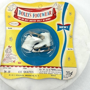 Op de afbeelding: Vintage verpakking met witte plastic schaatsen voor poppen. De geel en blauwe verpakking heeft de tekst "DOLLY'S FOOTWEAR" en "ICE SKATES". De verpakking is gemarkeerd met "SK-39" en "Non-Toxic".