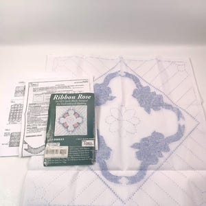 Puede incluir: Un conjunto de seis bloques de colcha blancos con diseños de rosas bordadas en azul. Los bloques están estampados para bordar y acolchar. El embalaje incluye el texto "Ribbon Rose" y "Set of 6 Quilt Blocks Stamped for Embroidery & Quilting".