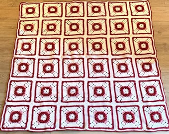 May include: A handmade crochet blanket featuring a repeating square pattern. Each square has a white background with a central burgundy flower design and burgundy border. The blanket is laid out on a wooden surface.