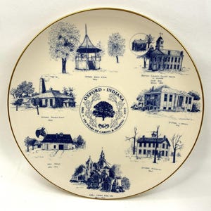 May include: A round ceramic commemorative plate with a gold rim, featuring blue ink illustrations of Oxford, Indiana landmarks. The center displays an oak tree emblem with the text Oxford, Indiana 1843-1993 150 Years of Caring & Sharing.