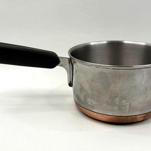 Puede incluir: Cacerola de acero inoxidable con base de cobre y asa negra. La olla pequeña tiene un solo mango largo y un cuerpo redondeado. La base de cobre añade un toque decorativo a la olla.
