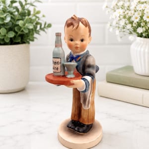 May include: A ceramic figurine of a young person dressed in a dark jacket, blue bow tie, and brown trousers, holding a round red tray. The tray carries a grey bottle labeled Rhein-Wine and a small grey goblet. A white cloth hangs from the arm.