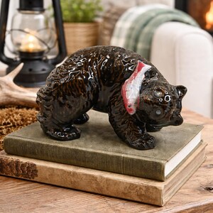 May include: A ceramic bear figurine with a glossy, dark brown finish, holding a red and white fish. The bear is posed on top of two vintage books with aged covers, suggesting a rustic or cabin-themed decor.