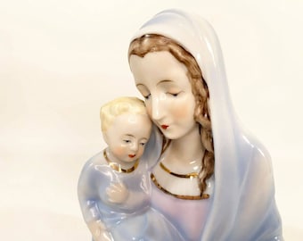 May include: A porcelain figurine of the Virgin Mary holding a baby Jesus. The figurine is painted in shades of blue and white, with gold accents. The Virgin Mary is wearing a blue robe and a white veil, and the baby Jesus is wearing a blue robe.