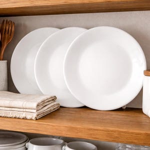 May include: A kitchen shelf displays three white plates, wooden utensils in a speckled ceramic container, folded beige linen napkins, a white jar with a wooden lid, garlic bulbs, and a glass bottle of oil.