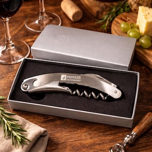May include: A stainless steel corkscrew with the text "PIONEER Investments" in a silver gift box. The image includes wine glasses with red wine, a cork, cheese, and grapes, suggesting a wine-tasting theme.