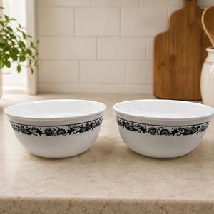 May include: Two white glass bowls featuring a continuous black floral pattern around the exterior rim. The bowls have a smooth, rounded shape and a glossy finish, showcasing a detailed botanical design in black ink against the white glass surface.