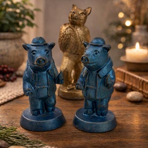 May include: Three decorative figurines: two blue pig statues wearing hats and jackets, and a gold cat statue. The pig statues are on round bases. The cat statue is standing in the background. The figurines are on a wooden surface.